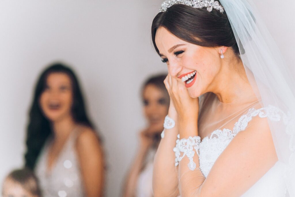 Profile view of bridge smiling in wedding dress adjusting earring with bridesmaids in background